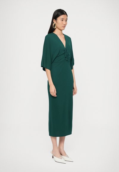 Πράσινο φόρεμα μέχρι το γόνατο με βαθύ V-λαιμόκοψη και λε�πτομέρειες με πιετάκια στη μέση. Διαθέτει φαρδιά μανίκια και εφαρμοστή σιλουέτα. Άσπρα mule με τακούνι.