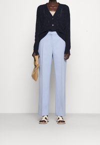 Personne portant un cardigan en tricot noir, un pantalon tailleur bleu clair, des sandales blanches, tenant un sac beige sur un fond uni.