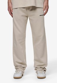 Pantalones de chándal beige con un corte relajado, hechos de una suave mezcla de algodón, con un pequeño logo negro 'PEGADOR' en el muslo izquierdo.
