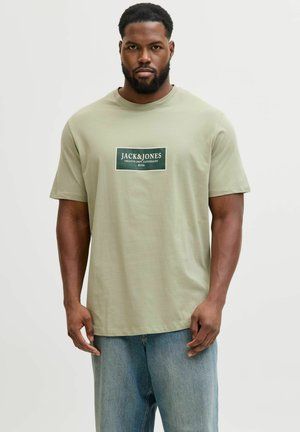 Mand med kort krøllet hår og skæg iført en lysegrøn Jack & Jones T-shirt og blå jeans stående mod en ensfarvet baggrund.