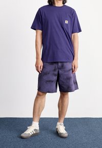 T-shirt di cotone viola con taschino sul petto, abbinata a pantaloni corti tie-dye viola scuro. Indossata con sneaker grigie e bianche e calze bianche.