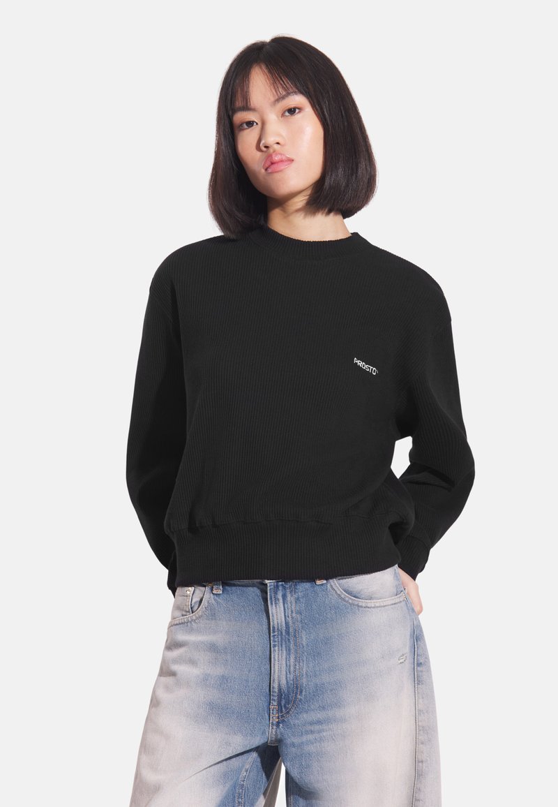 Czarna prążkowana bluza z okrągłym dekoltem i haftowanym logo. Noszona z wyblakłymi niebieskimi jeansami. Miękka w dotyku, o luźnym kroju.