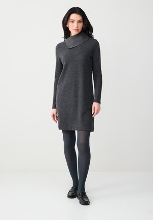 Femme portant une robe en maille à manches longues gris foncé avec un large col bénitier, des collants foncés et des mocassins noirs, debout devant un fond clair uni.