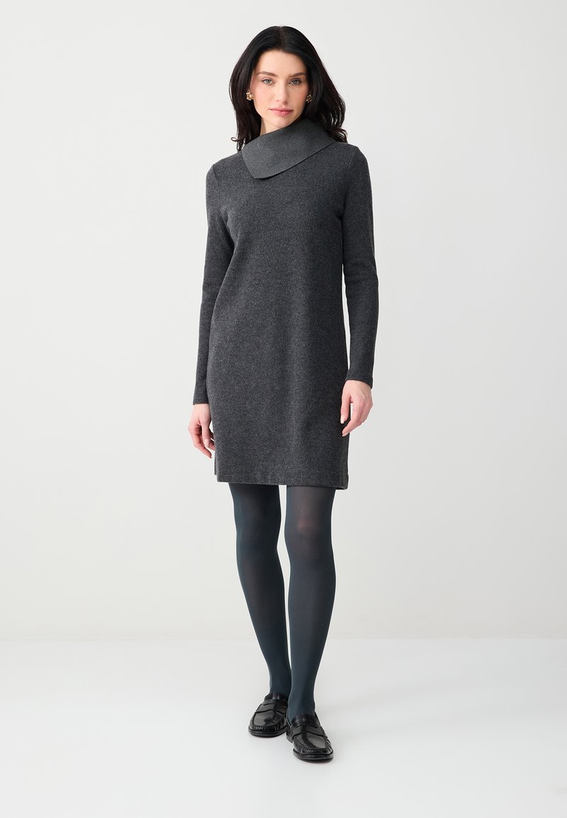 Femme portant une robe en maille à manches longues gris foncé avec un large col bénitier, des collants foncés et des mocassins noirs, debout devant un fond clair uni.