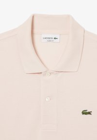 Pikétröja i ljus persikofärgad tyg, med klassisk krage, två knappar och en broderad grön krokodil-logotyp på vänster bröst.