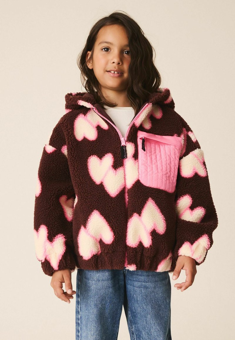 Enfant portant une veste polaire marron avec des motifs de cœurs roses et blancs et une poche poitrine matelassée rose, associée à un jean bleu.