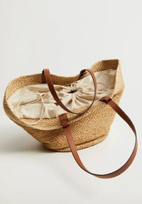 Woven straw tote with brown leather straps, featuring a beige drawstring fabric lining. Shallow, wide shape with a soft texture.