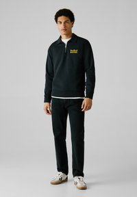 Zwarte zip-up sweatshirt met "Red Bull Racing" in het geel, gecombineerd met donkere jeans en witte sneakers. Glad materiaal, ontspannen pasvorm.