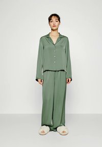 Grön satinpyjamas som består av en knäppt topp med krage och långa ärmar, tillsammans med löst sittande byxor och fluffiga vita tofflor.