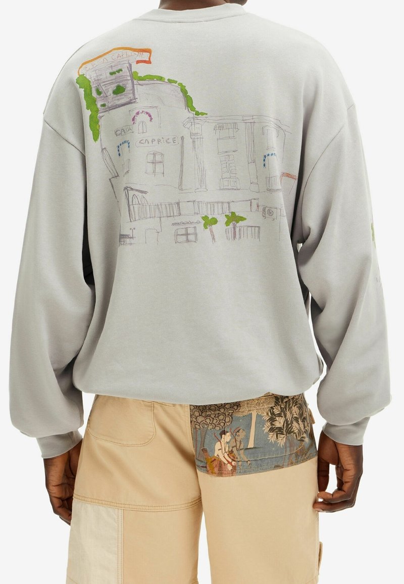 Sudadera gris claro de mangas largas, con coloridos diseños de bocetos de edificios y vegetación en la parte trasera. Combinada con pantalones cargo beige.