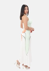 Femme dans une robe maxi sans manches vert pastel et blanche à fleurs avec une fente latérale, portant des sandales à talons blancs, se tient de côté en regardant l'appareil photo.