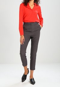 Blusa rossa a maniche lunghe con scollatura a V, abbinata a pantaloni tapered a fantasia bianca e nera. Completano l'outfit delle scarpe nere a punta senza lacci.