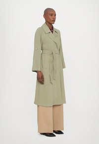 Trench coat verde chiaro di media lunghezza con cintura, maniche larghe e tasche frontali. Abbinato a pantaloni beige a gamba larga e scarpe nere.