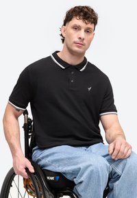 Άνδρας με κατσαρά μαλλιά που φοράει μαύρο πόλο και μπλε τζιν, κάθεται σε αναπηρικό καροτσάκι και κρατάει τον τροχό, ουδέτερη έκφραση, λευκό φόντο.