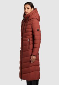 Rød foret jakke med høj krave og snøre, med chevron-quilting, en sidelomme med lynlås og et sort logo-patch på ærmet.