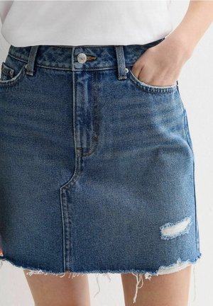 Mini-jupe en denim avec ourlet effiloché et patch usé, portée avec un haut blanc, main de la personne dans la poche avant du côté droit.