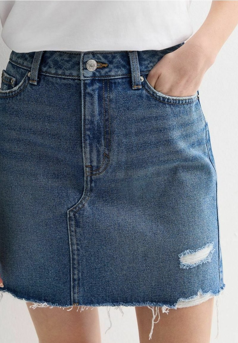 Mini-jupe en denim avec ourlet effiloché et patch usé, portée avec un haut blanc, main de la personne dans la poche avant du côté droit.