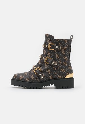 Bottine noire à la cheville avec un extérieur en cuir à motif, équipée d'une quincaillerie dorée, de trois boucles, de strass et d'une semelle noire épaisse.