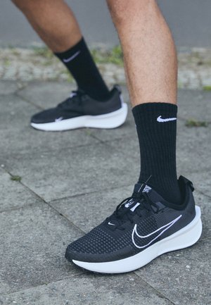 Osoba nosząca czarne buty do biegania Nike i czarne skarpetki Nike stoi na szarym, wyłożonym płytkami chodniku na zewnątrz.