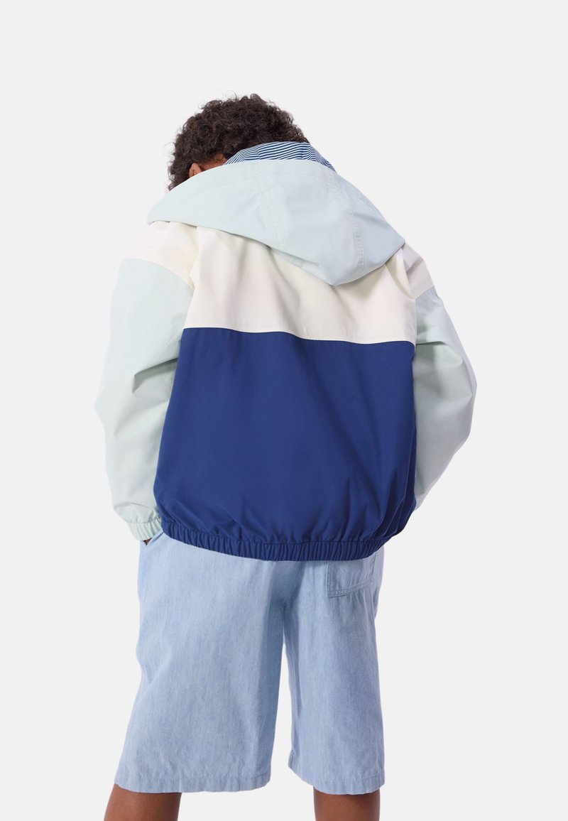 Giacca color block con sezioni blu chiaro, crema e navy. Presenta un cappuccio, un orlo elastico e un tessuto leggero; indossata con pantaloncini blu.