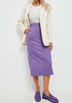 Kvinde iført hvid blazer, cremefarvet ribbet top, lilla midi-nederdel med lommer og matchende lilla loafers, der står op ad en hvid væg.