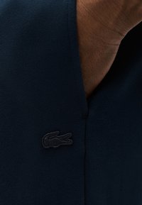 Χέρι με σκούρο δέρμα που εισέρχεται εν μέρει στην τσέπη από σκουρόχρωμο ναυτικό ύφασμα με ένα μικρό κεντητό λογότυπο κροκόδειλου κοντά στο άνοιγμα.