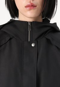 Czarna kurtka z kapturem z metalowym zamkiem z przodu, detal z zatrzaskiem oraz miękka, fakturowana tkanina. Posiada luźny krój w okolicy ramion.