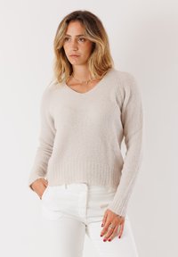 Maglione a maglia beige chiaro con scollatura a V, maniche lunghe e orlo a costine. Texture morbida e vestibilità rilassata, abbinato a pantaloni bianchi.