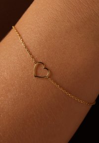 Pulsera de oro con un colgante en forma de corazón con un diseño abierto, enlazada por una delicada cadena, descansando sobre una superficie de piel texturizada.