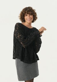 Top in pizzo nero con maniche lunghe, caratterizzato da una vestibilità ampia e da intricati motivi floreali. Abbinato a una gonna grigia. Tessuto leggero visibile.