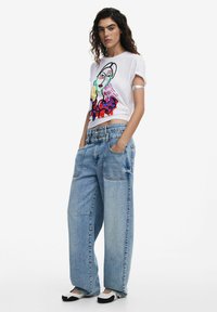 Weißes Grafik-T-Shirt mit einer bunten Gesichtillustration, kombiniert mit hellblauen, übergroßen Jeanshosen und schwarzen und beigen Slippern.