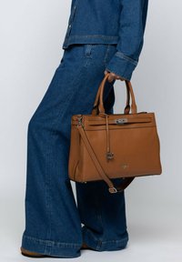 Brown leather handbag with dual top handles, adjustable crossbody strap, zip closure, and a textured finish. Features silver-tone accents.