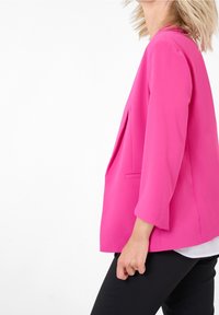 Kvinna i en ljusrosa blazer, vit skjorta och svarta byxor, stående på sidan med handen avslappnat vid sidan.