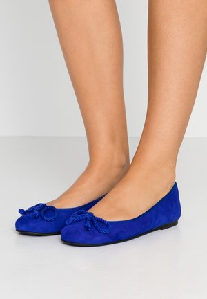 Pieds portant des ballerines en daim bleu vif avec des bouts arrondis et des nœuds décoratifs en corde sur un sol gris clair.