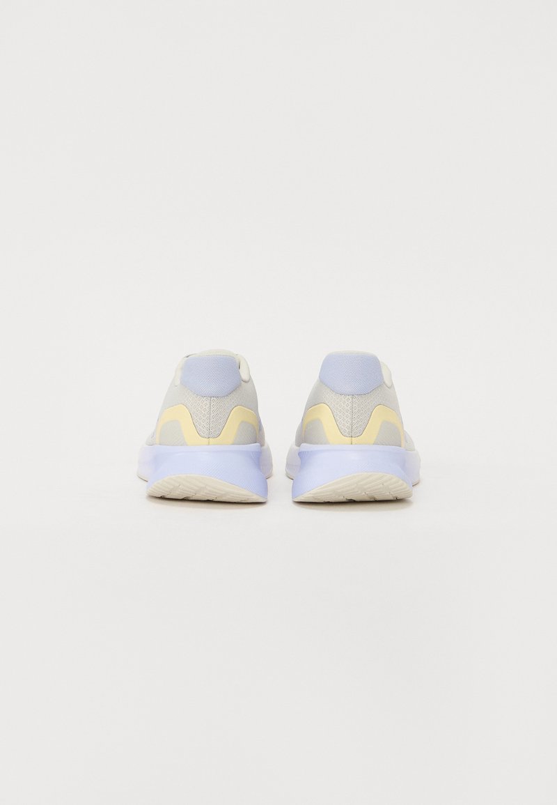 Zapatillas deportivas de color gris claro con una textura suave, con acentos en amarillo y lavanda, suela acolchada y un diseño trasero liso.