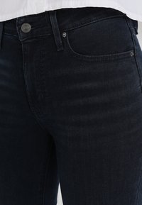 Vaqueros de mezclilla oscura con un corte ajustado. Las características incluyen un bolsillo frontal, cierre con botón de metal y sutiles detalles de costura. Textura suave.