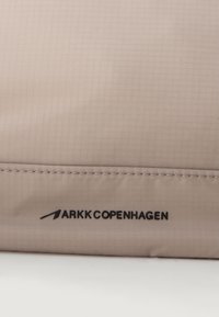 Tejido beige suave con un patrón a cuadros, que presenta la marca "ARKK COPENHAGEN" en negro y costuras visibles.