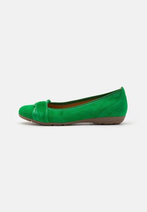 Chaussure plate en daim vert avec un nœud décoratif sur l’orteil et une semelle en caoutchouc marron, vue de profil sur fond blanc.