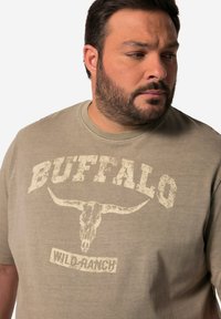 Tričko Buffalo Wild Ranch v světle hnědé barvě s kulatým výstřihem. Obsahuje vybledlý grafický motiv lebky skotu a tučné písmo. Měkká textura.