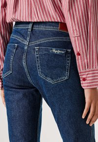 Tumši zili džinsu bikses ar pieguļošu dizainu, vidējas augstuma jostasvietu, divām aizmugurējām kabatām un smalkām šuvēm, kopā ar sarkanu svītrainu kreklu.