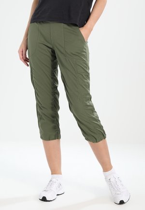 Person trägt olivgrüne, kurz geschnittene Hose mit Fronttaschen, elastischen Bündchen, schwarzes Oberteil und weiße Sportschuhe, steht vor weißem Hintergrund.