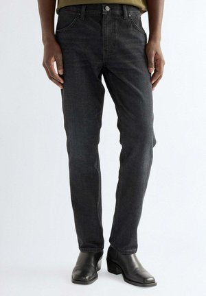 Mann trägt schwarze Slim-Fit-Jeans und schwarze Leder-Stiefeletten, steht vor einem schlichten hellen Hintergrund.