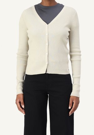 Person trägt eine eng anliegende beige gerippte Strickjacke mit Knöpfen über einem dunkelgrauen Rundhalsausschnitt-Shirt und schwarzen Hosen mit weitem Bein.