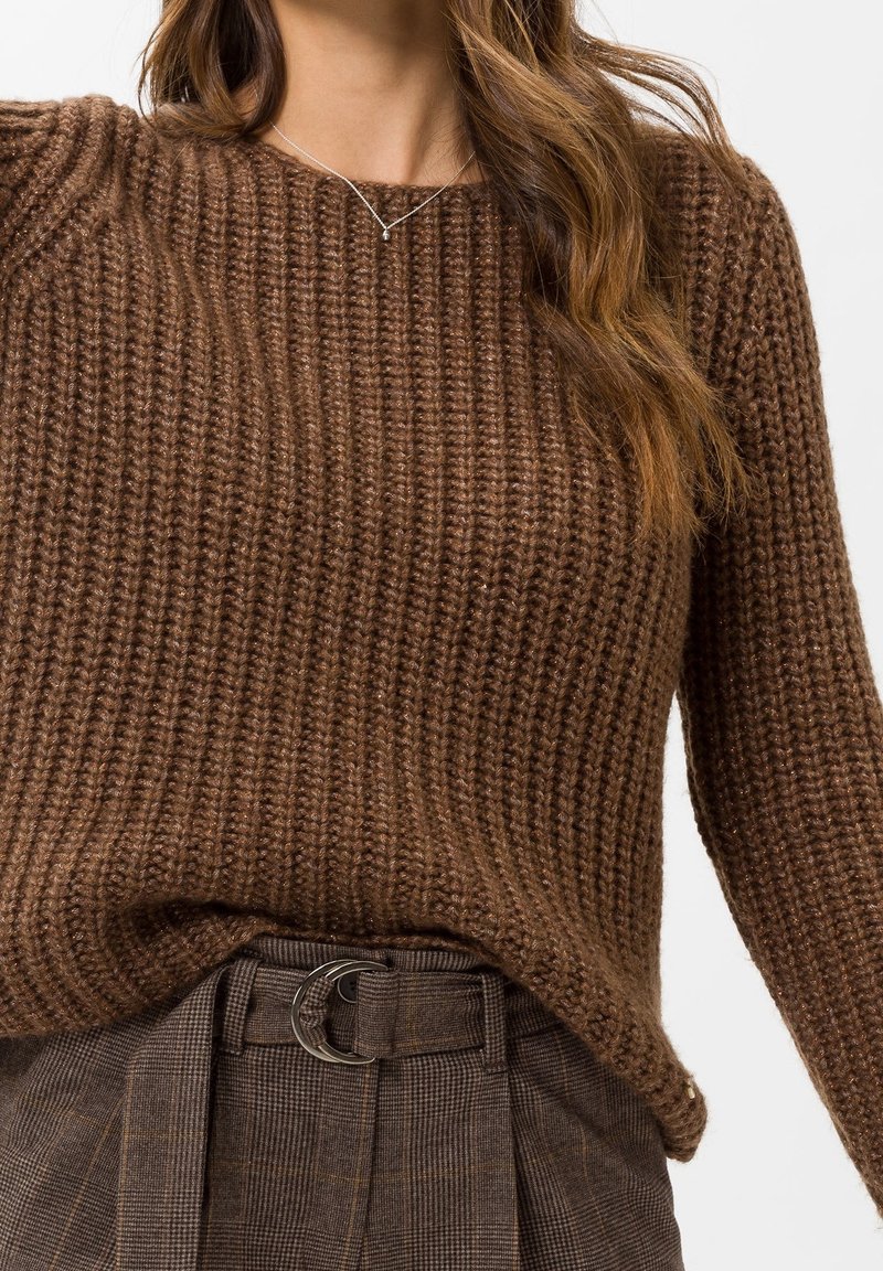 Pull en maille marron avec une texture côtelée, présentant une coupe ample et un décolleté large, associé à un pantalon à carreaux marron et une boucle de ceinture.