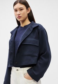 Femeie purtând o jachetă scurtă bleumarin peste un pulover închis la culoare și pantaloni deschiși la culoare, cu talie înaltă, cu o mână în buzunar, pe un fundal simplu.