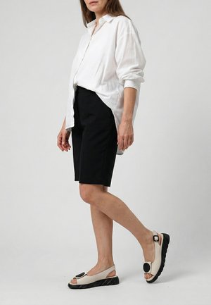 Vrouw draagt een losse witte blouse met knopen, zwarte knielange korte broek en beige slingback sandalen met zwarte zolen en ronde zwarte accenten.