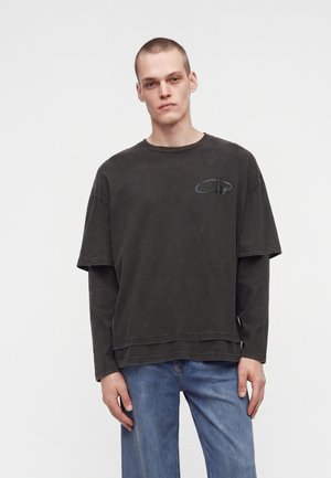 Ung mand med kort hår iført en lagdelt mørkegrå langærmet trøje og blå jeans, stående mod en ensartet lys baggrund.