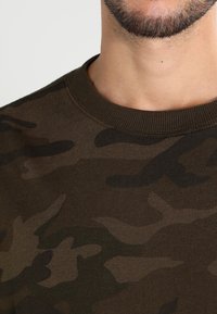 Felpa mimetica verde scuro con colletto rotondo, girocollo e texture morbida, che presenta diverse tonalità di verde e marrone.