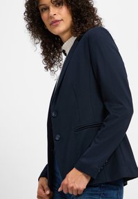 Marineblazer aus strukturiertem Stoff, mit zwei blauen Knöpfen, mitrevers und einer einzelnen Brusttasche. Körpernahe Passform mit Seitentaschen.