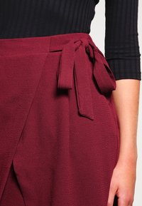 Burgundy omslagskjol i en texturerad tyg, med en knuten midjebanddetalj. Tyget verkar mjukt och svävande, vilket förhöjer designen.